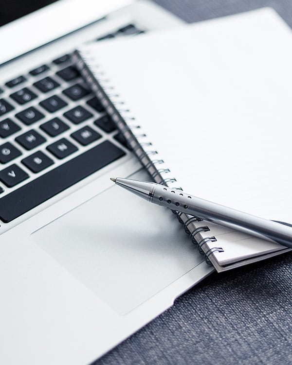 Pen resting ontop of a blank notepad and an open laptop